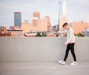 teen boy walks parallel to camera looking down along a parking garage rooftop wall with the Oklahoma City skyline behind him