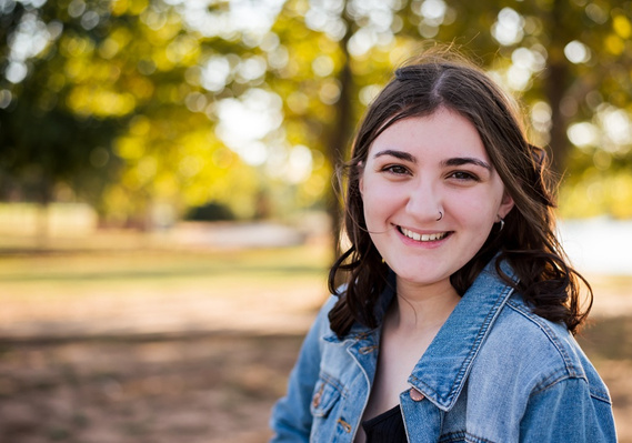 A high school senior girl stands and smiles in front of the trees and pond in Shannon Springs Park in Chickasha, Oklahoma 