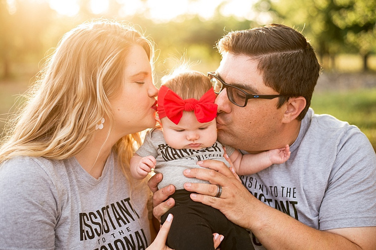 Mom and Dad kiss their baby girl on the cheeks in a park.