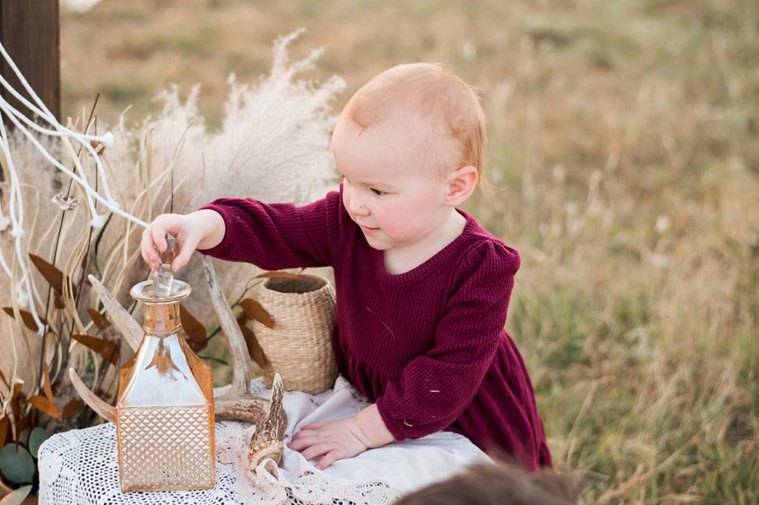 A toddler girl lifts the lid off a decanter at a boho photo set in a field at a family photo session in Tuttle, Oklahoma