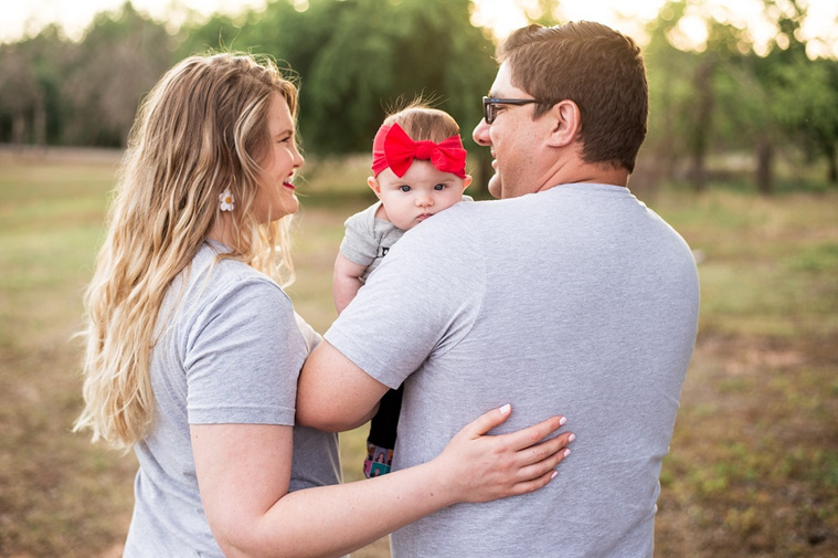 Mom and Dad hold their baby girl in the park while she looks over her dad's shoulder.