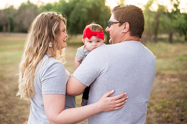 Mom and Dad hold their baby girl in the park while she looks over her dad's shoulder.