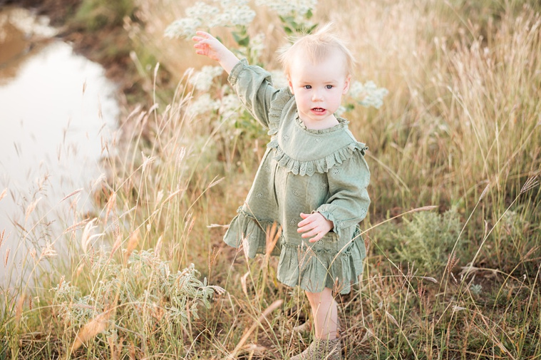 A toddler girl in a green dress and leopard booties stands and points to a pond in a field at a family photo session in Tuttle, Oklahoma
