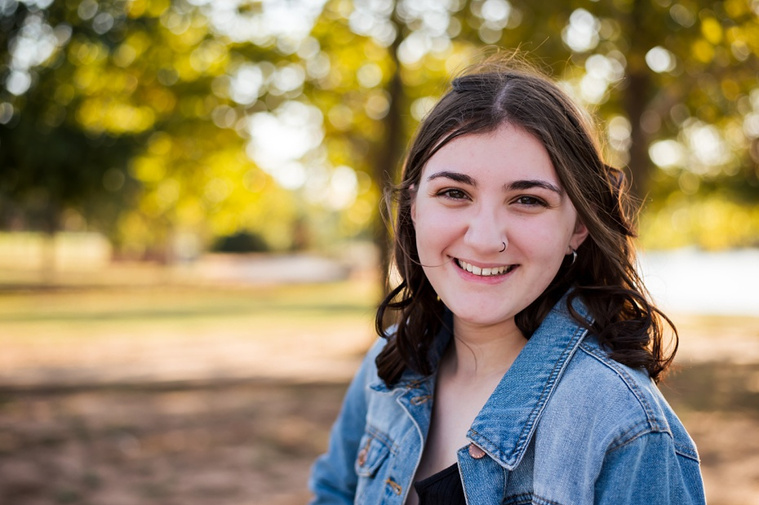 A high school senior girl stands and smiles in front of the trees and pond in Shannon Springs Park in Chickasha, Oklahoma 