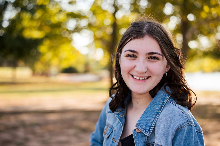 A high school senior girl stands and smiles in front of the trees and pond in Shannon Springs Park in Chickasha, Oklahoma 