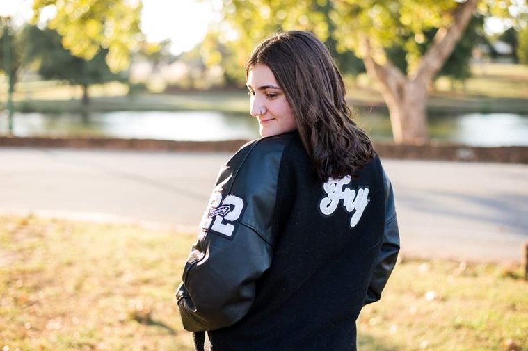A high school senior girl wearing her letterman jacket stands and smiles in front of the trees and pond in Shannon Springs Park in Chickasha, Oklahoma 