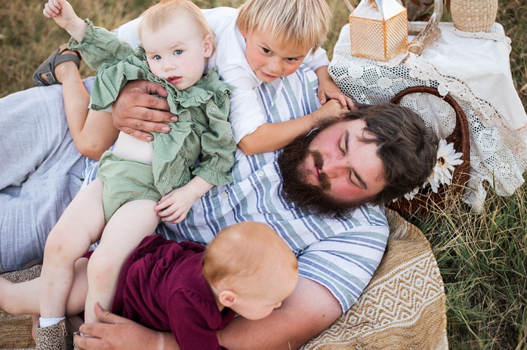 A dad plays on the ground with his three young children in a field at a family photo session in Tuttle, Oklahoma