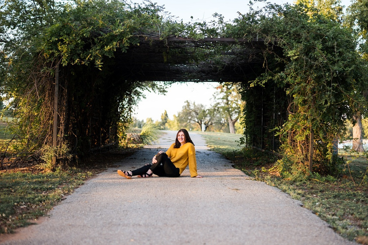 A high school senior girl sits on a path in front of an ivy covered structure in Shannon Springs Park in Chickasha, Oklahoma 