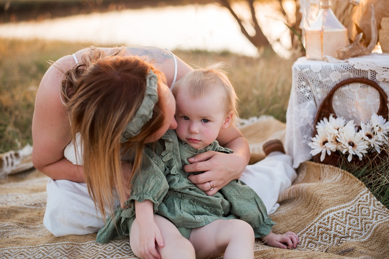 A mom kisses her toddler daughters cheek sitting on a rug in a field at a family photo session in Tuttle, Oklahoma