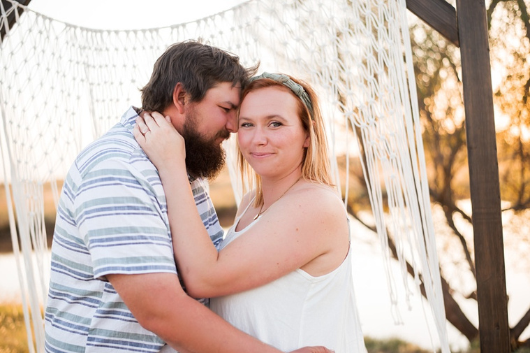 a husband and wife snuggle in front of a macrame curtain in a field at a family photo session in Tuttle, Oklahoma