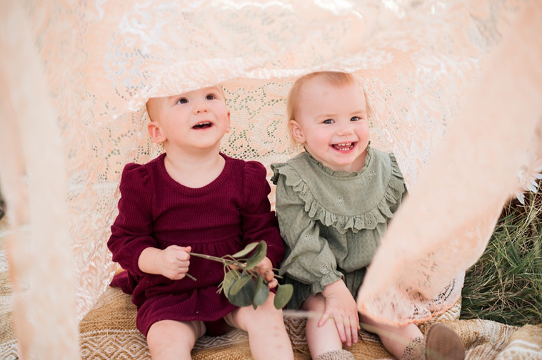 Two twin toddler girls play peek-a-boo and laugh and smile in a field at a family photo session in Tuttle, Oklahoma