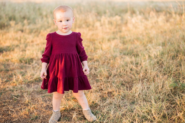A toddler girl in a red dress and leopard booties stands in a field at a family photo session in Tuttle, Oklahoma