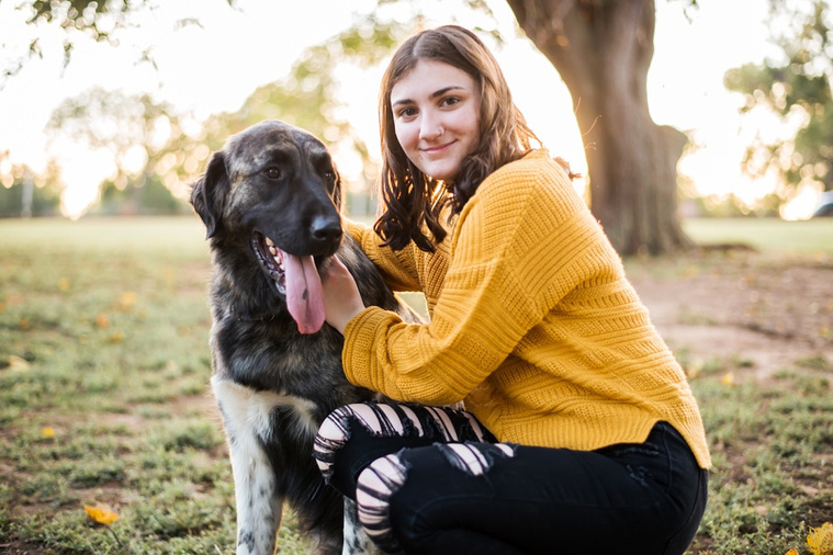 A high school senior girl crouches and smiles with her large dog in front of the trees and pond in Shannon Springs Park in Chickasha, Oklahoma 