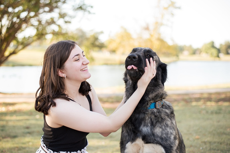 A high school senior girl sits in the grass with her dog and laughs as his tongue pokes out in front of the trees and pond in Shannon Springs Park in Chickasha, Oklahoma 