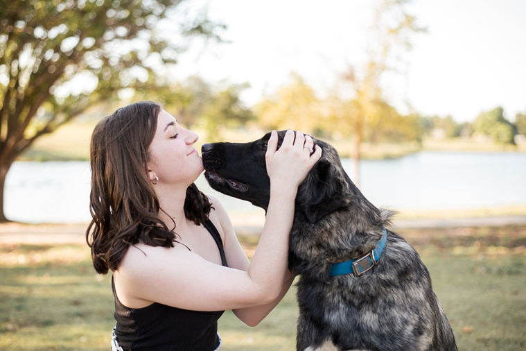 A high school senior girl sits on the grass with her large dog and nuzzles him in front of the trees and pond in Shannon Springs Park in Chickasha, Oklahoma 