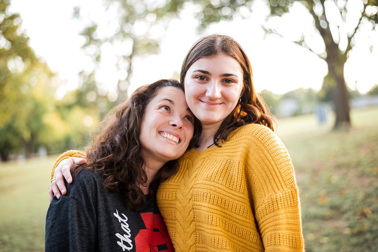 A high school senior girl stands and smiles with her mom in front of the trees and pond in Shannon Springs Park in Chickasha, Oklahoma 