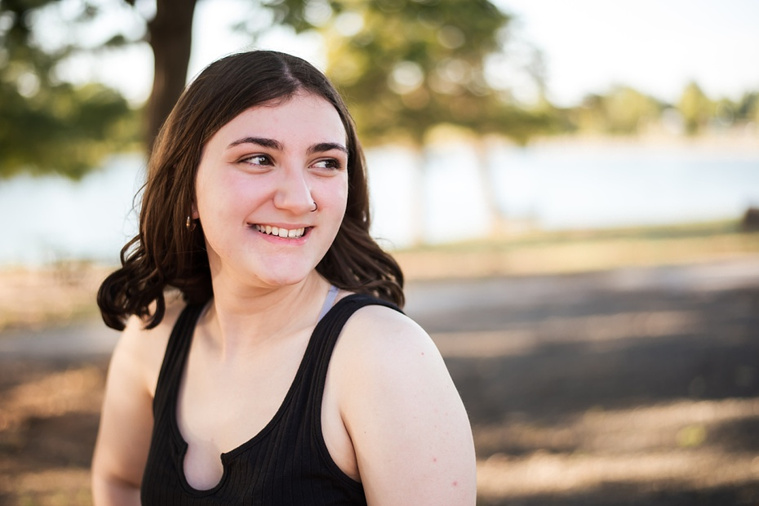A high school senior girl stands and smiles in front of the trees and pond in Shannon Springs Park in Chickasha, Oklahoma 