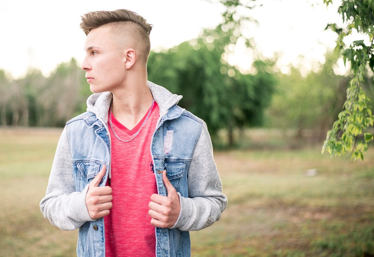 Teen boy dressed modern stands looking off his side with hands on jacket in a park in Oklahoma