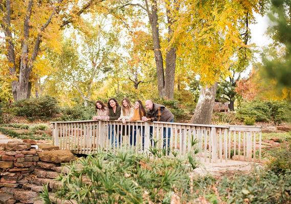 A mom and dad and 3 teen daughters stand on a wooden footbridge and smile together with fall color all around them at Will Rogers Gardens in Oklahoma City, Oklahoma