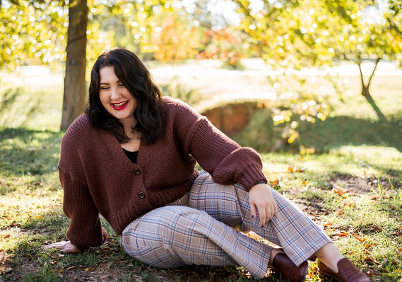 A Senior girl sits in the grass and laughs in front of fall color trees with sunlight behind her at Will Rogers Gardens in Oklahoma City Oklahoma