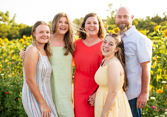 A mom and dad with three teen daughters stand together and smile in a flower field of sunflowers near Oklahoma City, Oklahoma.