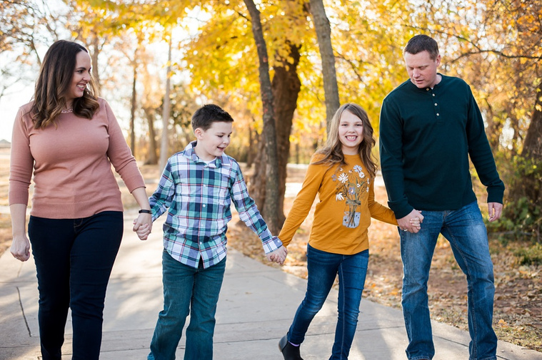 a family of four with two young children hold hands and walk on a sidewalk in a park in Moore, Oklahoma with golden leaves on the trees behind them at a family photo session