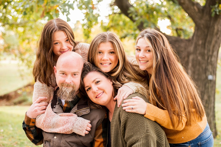 A mom and dad with 3 teen daughters sit and smile together on a bench with fall color around them at Will Rogers Gardens in Oklahoma City, Oklahoma