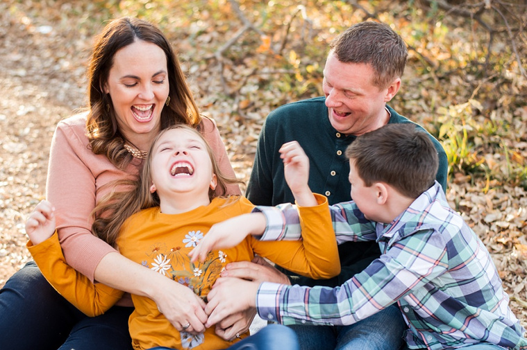 a mom and dad sit on the ground in the woods with their son and daughter. Everyone is tickling the girl and they all are laughing. 