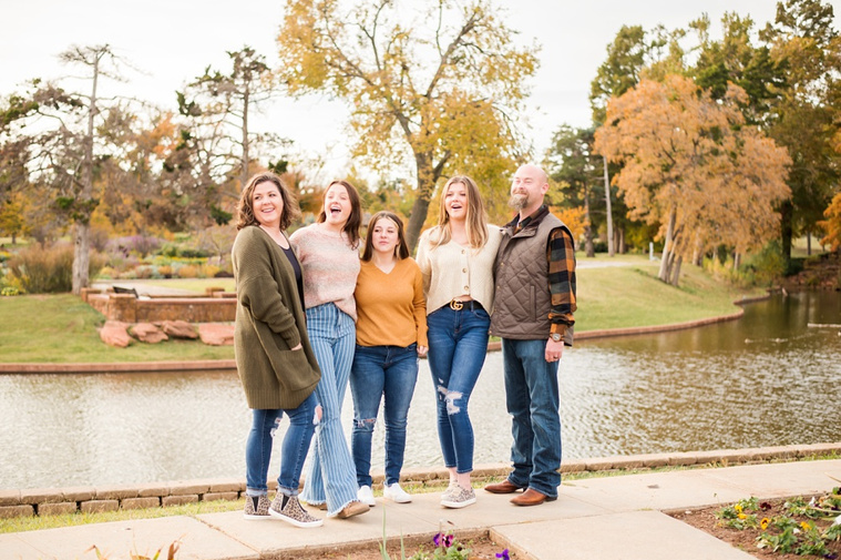 A mom and dad with 3 teen daughters stand and smile together on a sidewalk with fall color around them and a pond behind them at Will Rogers Gardens in Oklahoma City, Oklahoma