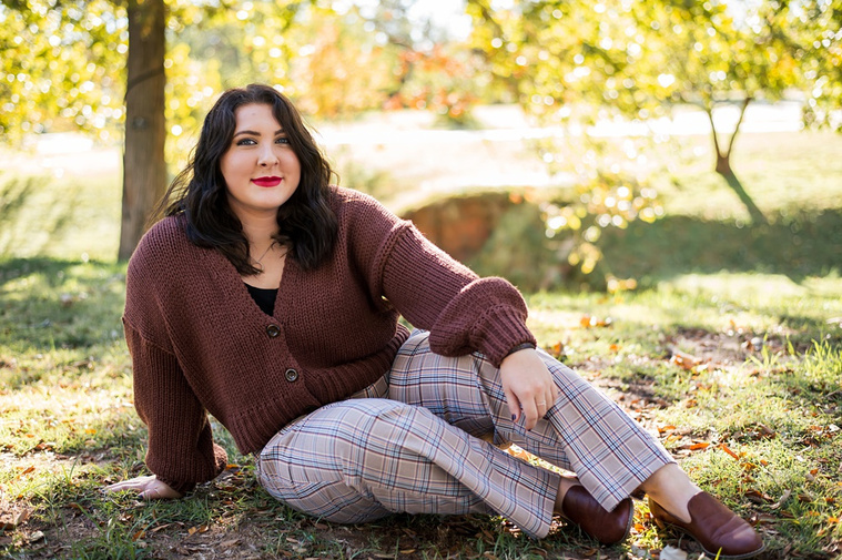 Professional photo of a senior girl sitting in the grass and smiling with colorful fall trees behind her at Will Rogers Gardens in Oklahoma City, Oklahoma
