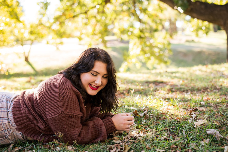 Professional photo of a senior girl laying on her stomach in the grass, looking downward and laughing, with colorful fall trees behind her at Will Rogers Gardens in Oklahoma City, Oklahoma