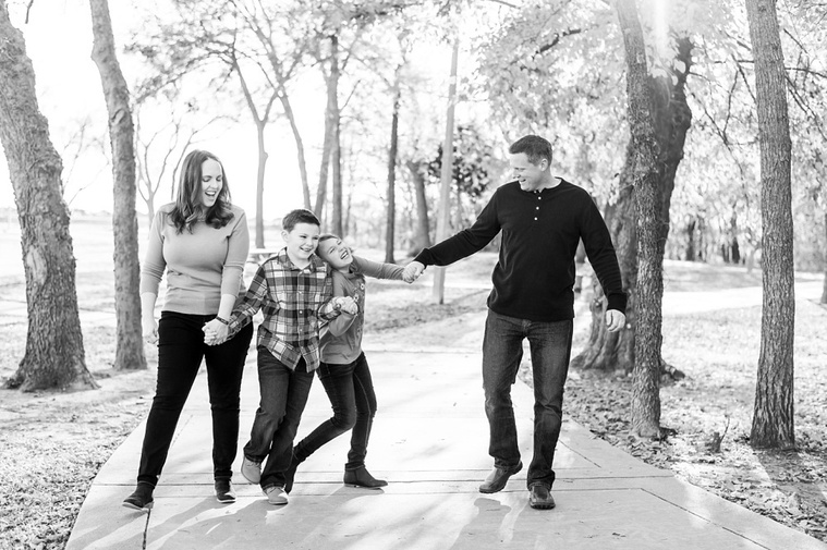 black and white photo of a mom and dad holding hands, walking and laughing on a sidewalk with their son and daughter at a family photo session in Oklahoma at a park in the fall