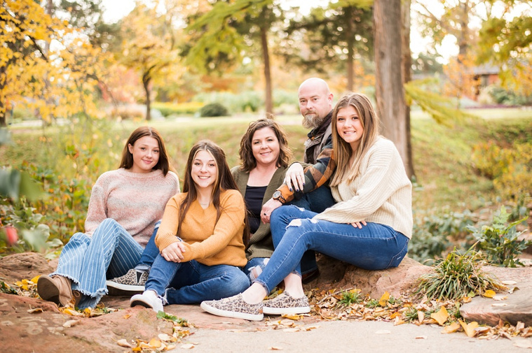 A mom and dad with 3 teen daughters sit and smile together on the rocks with fall color around them at Will Rogers Gardens in Oklahoma City, Oklahoma