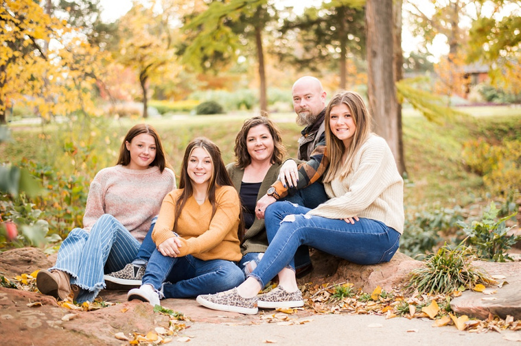 A mom and dad with 3 teen daughters sit and smile together on the rocks with fall color around them at Will Rogers Gardens in Oklahoma City, Oklahoma