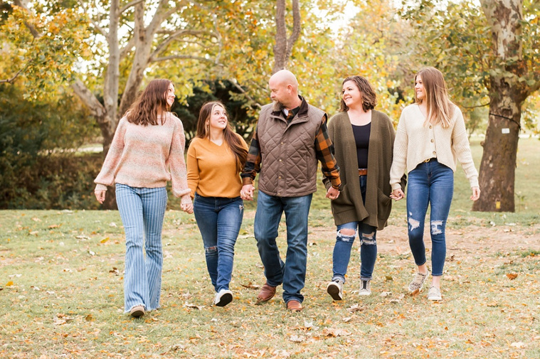 A mom and dad with 3 teen daughters walk towards camera and smile together with fall color around them at Will Rogers Gardens in Oklahoma City, Oklahoma