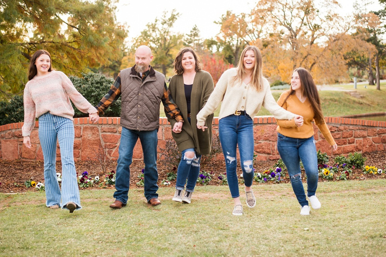 A mom and dad with 3 teen daughters walk towards camera, laughing together with flowers and a rock wall behind them, with fall color around them at Will Rogers Gardens in Oklahoma City, Oklahoma