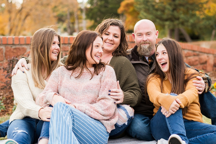 A mom and dad with 3 teen daughters sit and laugh together on the grass with flowers and a rock wall behind them and with fall color around them at Will Rogers Gardens in Oklahoma City, Oklahoma