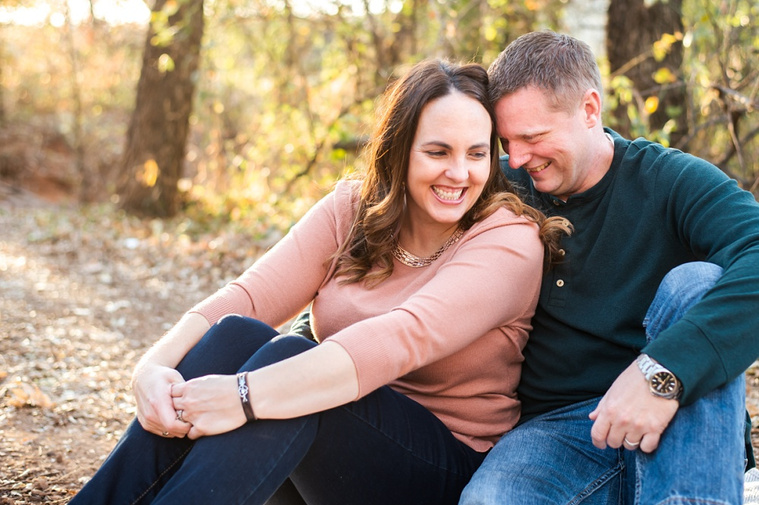 a husband and wife sit together and cuddle and laugh in the woods with fall color around them at a family photo session in Moore, Oklahoma
