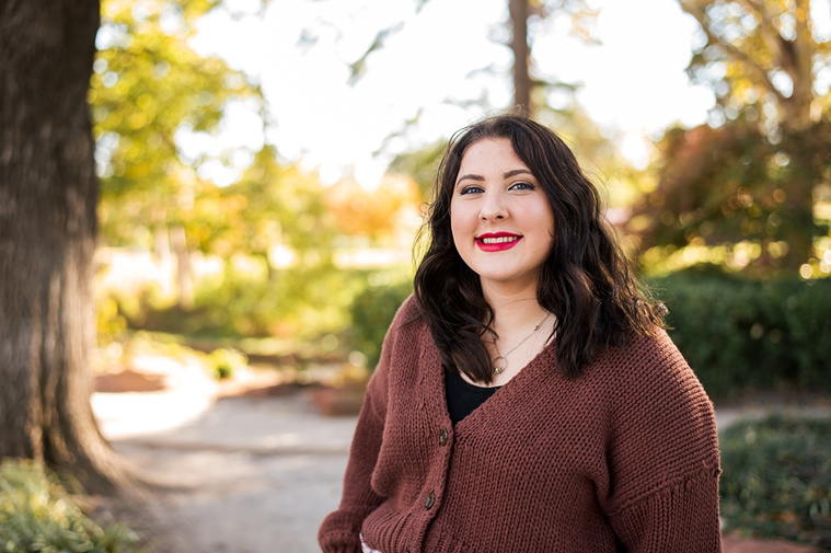 Professional photo of a senior girl standing on a path and smiling with colorful fall trees behind her at Will Rogers Gardens in Oklahoma City, Oklahoma