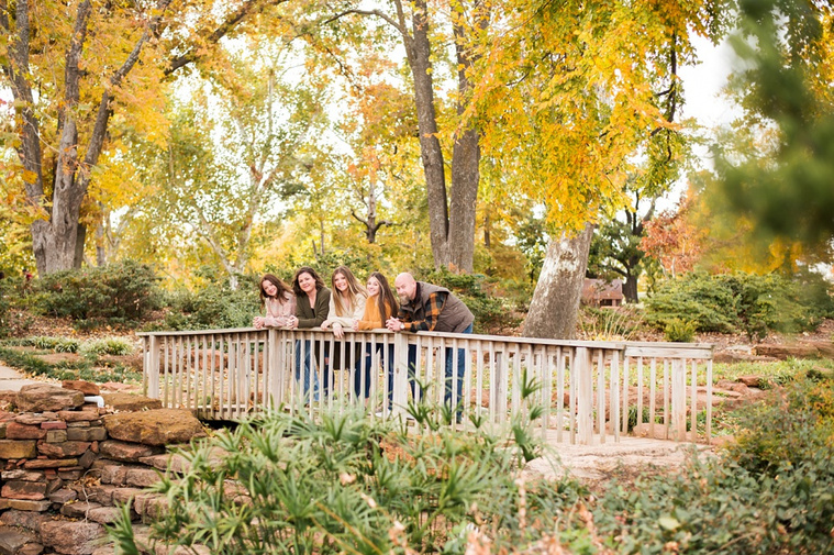 A mom and dad with 3 teen daughters stand on a wooden footbridge and smile together with fall color around them at Will Rogers Gardens in Oklahoma City, Oklahoma