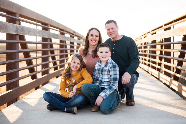 a mom, dad, young son and daughter sit together in the middle of a foot bridge at a park in Moore, Oklahoma at a family photo session