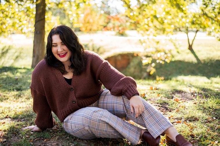 Professional photo of a senior girl sitting in the grass and laughing with colorful fall trees behind her at Will Rogers Gardens in Oklahoma City, Oklahoma