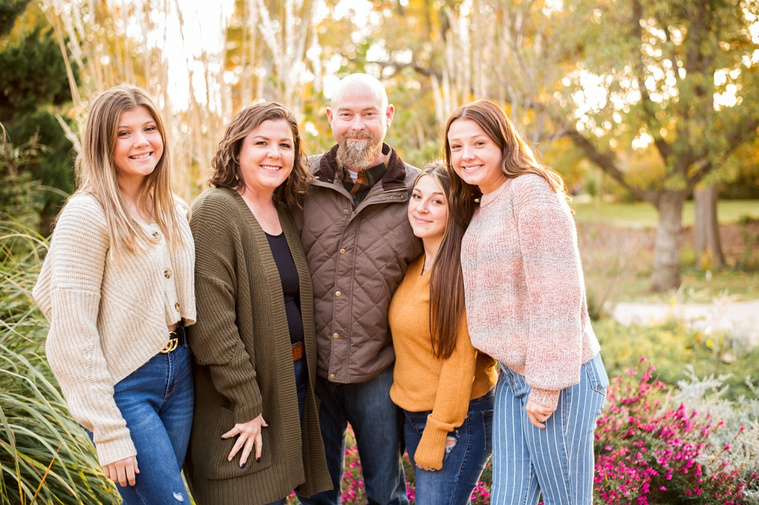 A mom and dad with 3 teen daughters stand and smile together in front of flowers with fall color around them at Will Rogers Gardens in Oklahoma City, Oklahoma