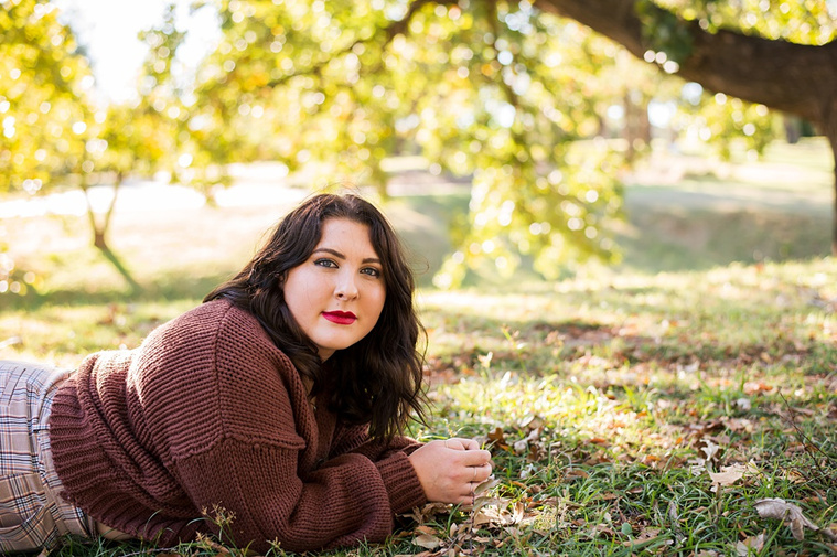Professional photo of a senior girl sitting in the grass and looking softly at the camera with colorful fall trees behind her at Will Rogers Gardens in Oklahoma City, Oklahoma
