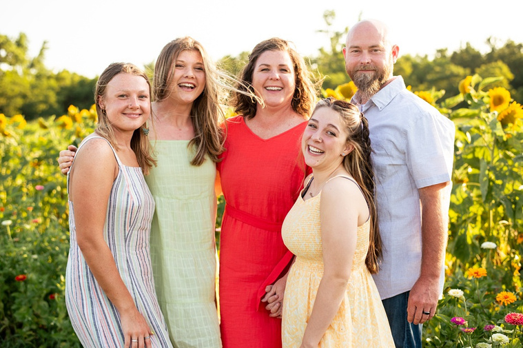 A mom and dad with three teen daughters stand together and smile in a field of sunflowers in Oklahoma.