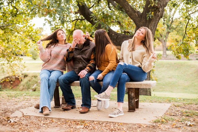 A dad with 3 teen daughters sit on a bench, dad has head in hands, teen girls are primping around him in a funny way, with fall color around them at Will Rogers Gardens in Oklahoma City, Oklahoma
