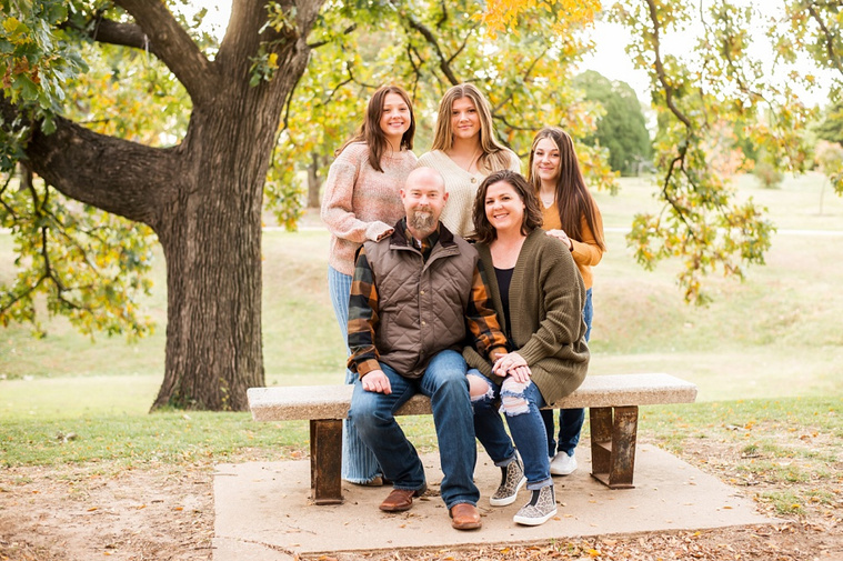 A mom and dad with 3 teen daughters sit and smile together on a bench with fall color around them at Will Rogers Gardens in Oklahoma City, Oklahoma