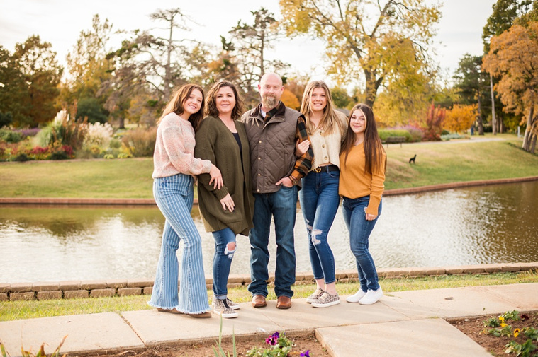 A mom and dad with 3 teen daughters stand on a sidewalk and smile together  with fall color around them and a pond behind them at Will Rogers Gardens in Oklahoma City, Oklahoma