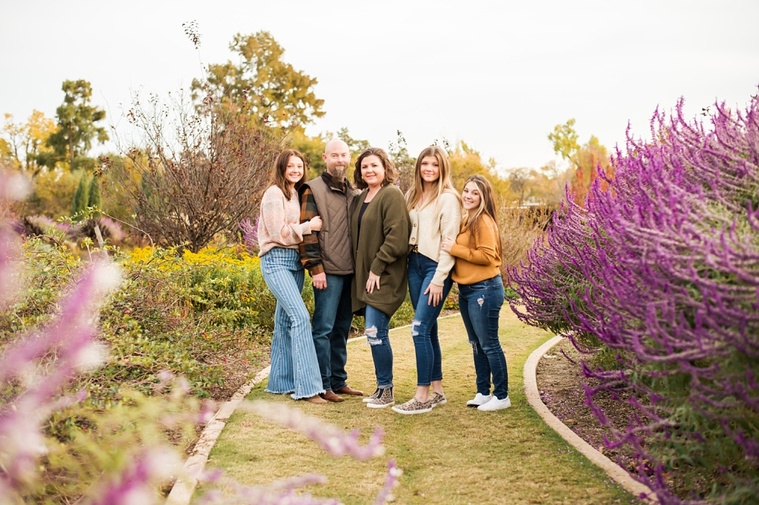 A mom and dad with 3 teen daughters stand and smile together on a path with purple flowers around them at Will Rogers Gardens in Oklahoma City, Oklahoma