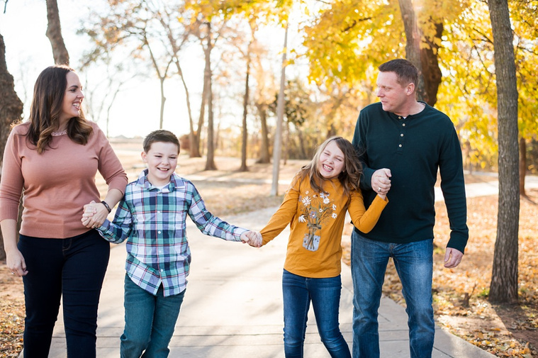 a family of four with two young children hold hands and walk on a sidewalk in a park in Moore, Oklahoma with golden leaves on the trees behind them at a family photo session